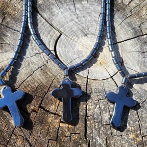 May include: Three black hematite cross necklaces on a wooden surface. Each necklace is made of small, round beads and features a large, flat cross pendant.