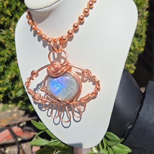 Op de afbeelding: Een handgemaakte koperdraad hangerketting met een ronde, iriserende edelsteen. De hanger is ingewikkeld bedraad in een oogachtig ontwerp, hangend aan een koperen kralenketting. De ketting wordt getoond op een witte buste tegen een achtergrond van groen.