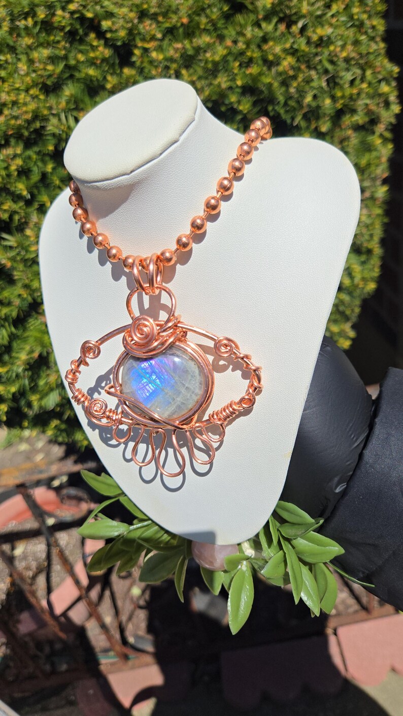 Op de afbeelding: Een handgemaakte koperdraad hangerketting met een ronde, iriserende edelsteen. De hanger is ingewikkeld bedraad in een oogachtig ontwerp, hangend aan een koperen kralenketting. De ketting wordt getoond op een witte buste tegen een achtergrond van groen.