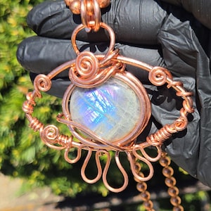 Op de afbeelding: Een handgemaakte koperdraad hangerketting met een grote, iriserende, ronde edelsteen. De hanger heeft een oogachtig ontwerp met spiraal- en lusdraaddetails. De ketting bevat een koperen ketting en kralenaccenten.
