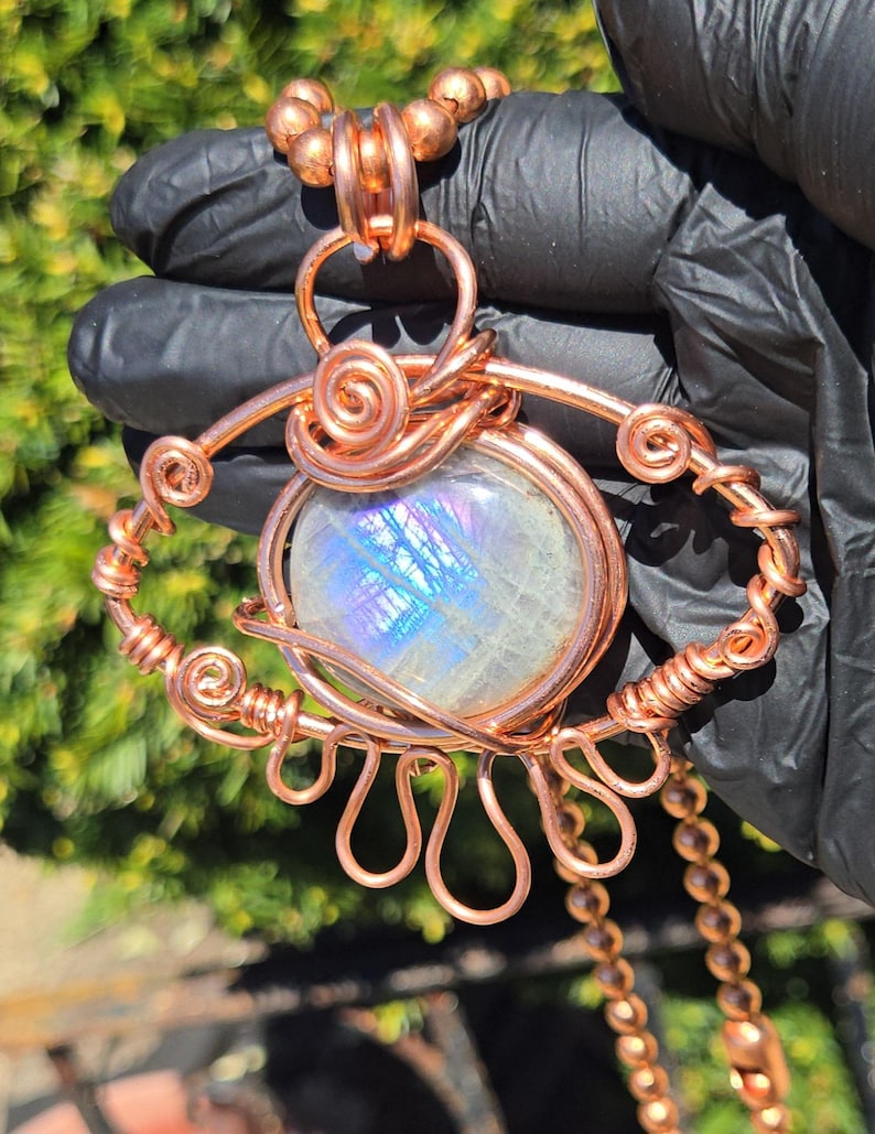 Op de afbeelding: Een handgemaakte koperdraad hangerketting met een grote, iriserende, ronde edelsteen. De hanger heeft een oogachtig ontwerp met spiraal- en lusdraaddetails. De ketting bevat een koperen ketting en kralenaccenten.
