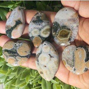 May include: A collection of six polished ocean jasper pendants in various shapes: teardrop, oval, and heart. The pendants feature a mix of colors including beige, green, and white, with unique patterns. Each pendant has a silver-colored bail for hanging.