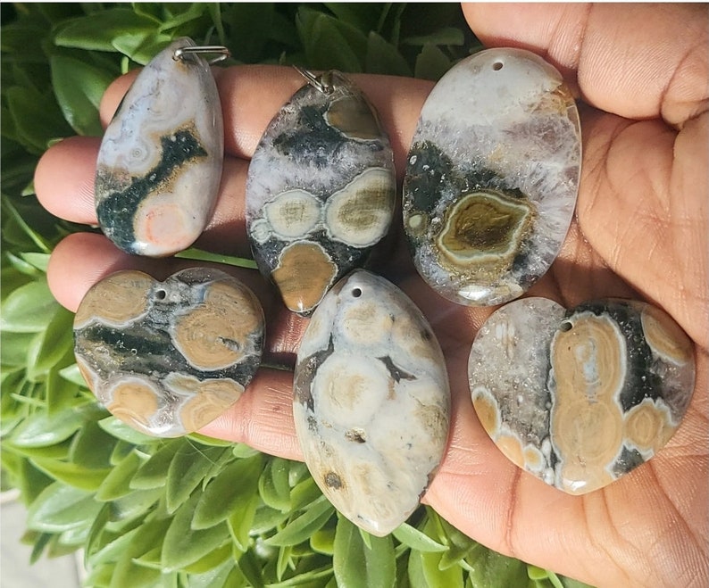May include: A collection of six polished ocean jasper pendants in various shapes: teardrop, oval, and heart. The pendants feature a mix of colors including beige, green, and white, with unique patterns. Each pendant has a silver-colored bail for hanging.