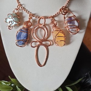 May include: A copper-toned ball chain necklace featuring five pendants. The center pendant is a wire-formed symbol, flanked by four wire-wrapped stones in light blue, dark blue, yellow, and purple. The metal components have a metallic copper finish.