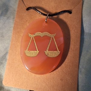 May include: A pink stone pendant necklace with a gold-colored zodiac symbol of Libra, the scales.