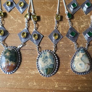 May include: A collection of gemstone necklaces and matching earrings. The necklaces feature oval and teardrop-shaped pendants with intricate patterns in shades of green, brown, and cream. The earrings and necklace chains are silver-toned, with yellow and green accent stones.