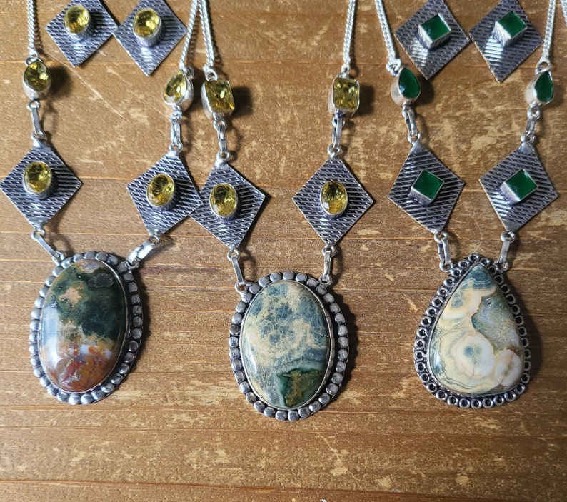 May include: A collection of gemstone necklaces and matching earrings. The necklaces feature oval and teardrop-shaped pendants with intricate patterns in shades of green, brown, and cream. The earrings and necklace chains are silver-toned, with yellow and green accent stones.