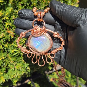 Op de afbeelding: Een handgemaakte koperdraad hangerketting met een grote, iriserende, ovaalvormige steen. De steen is gezet in een decoratief draadframe met wervelende en geluste ontwerpen. De ketting wordt vastgehouden door een koperen ketting.