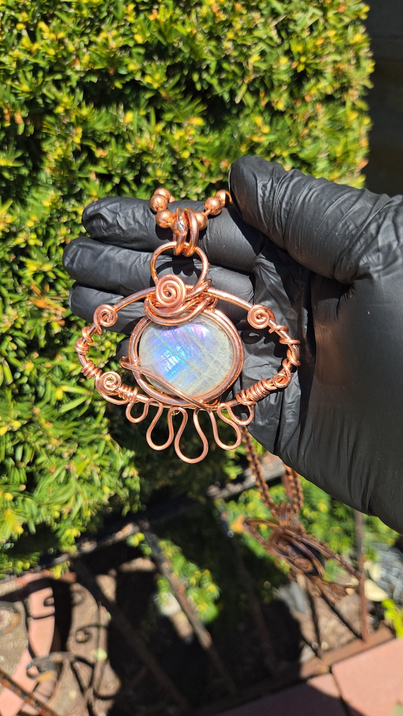 Op de afbeelding: Een handgemaakte koperdraad hangerketting met een grote, iriserende, ovaalvormige steen. De steen is gezet in een decoratief draadframe met wervelende en geluste ontwerpen. De ketting wordt vastgehouden door een koperen ketting.