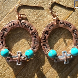 May include: Copper hoop earrings with the inscription "All things are possible. Matt 19:26." The earrings feature turquoise beads and a cross charm. The earrings are on a wooden surface.