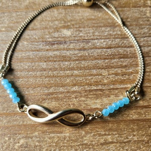 May include: A gold infinity charm bracelet with a sliding knot closure. The bracelet is adorned with light blue beads.