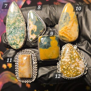 May include: A collection of gemstone rings with silver settings. The stones feature various patterns and colors, including green, yellow, and brown. The rings are displayed on a black surface, showcasing their unique designs and textures.