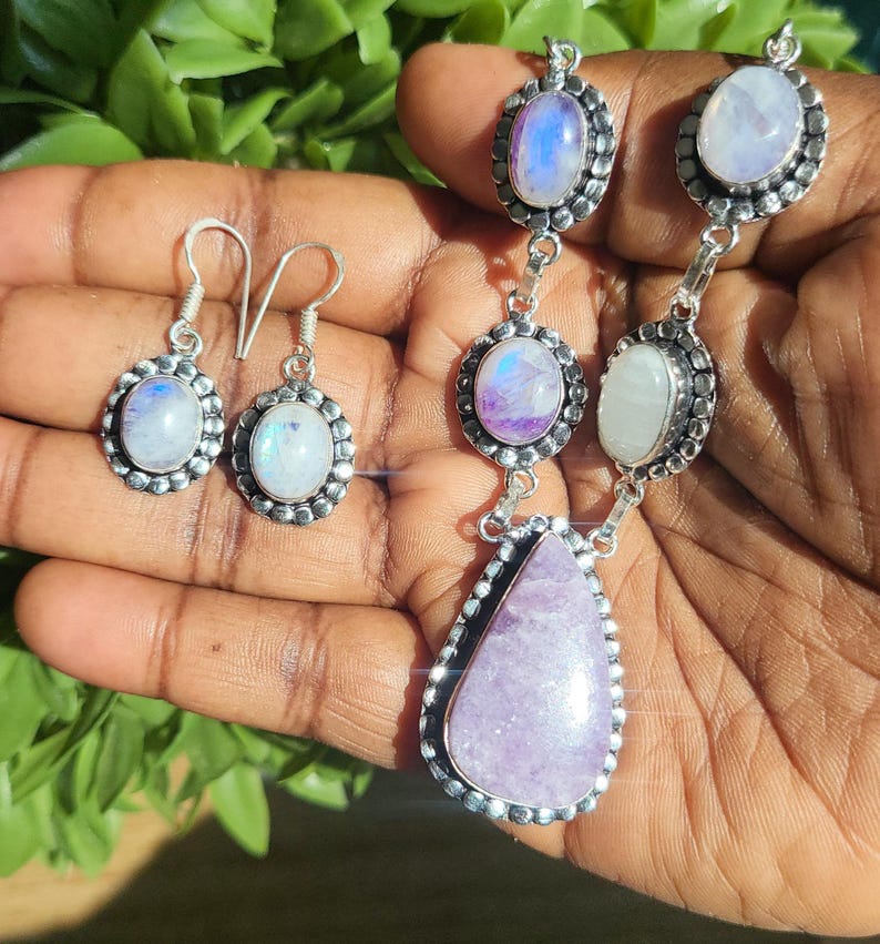 Puede incluir: Un conjunto de collar y pendientes de plata con piedras preciosas de piedra lunar y amatista. El collar presenta un colgante de amatista en forma de l&aacute;grima y tres piedras de luna redondas. Los pendientes son aros de plata con piedras de luna redondas.