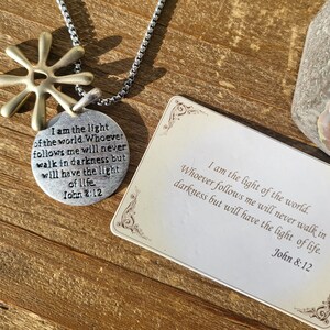 May include: A silver necklace with a round pendant featuring the text "I am the light of the world. Whoever follows me will never walk in darkness but will have the light of life. John 2:12". The pendant is attached to a silver chain with a gold star charm.