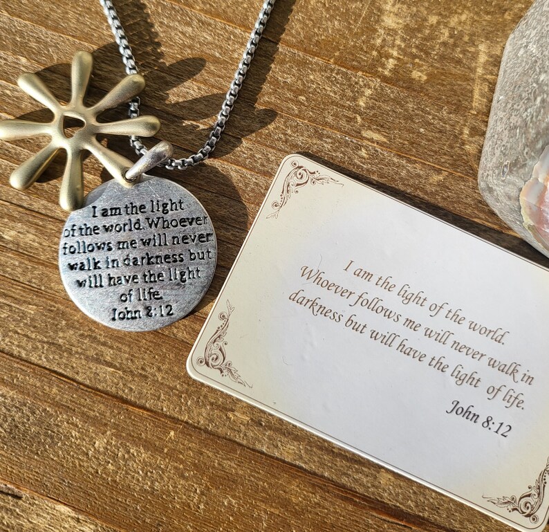 May include: A silver necklace with a round pendant featuring the text "I am the light of the world. Whoever follows me will never walk in darkness but will have the light of life. John 2:12". The pendant is attached to a silver chain with a gold star charm.