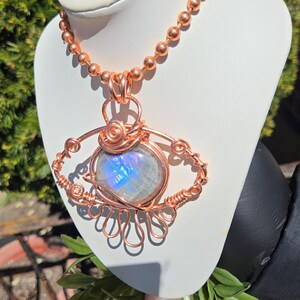 Op de afbeelding: Een handgemaakte koperdraad hanger ketting met een grote, iriserende maansteen. De maansteen is gezet in een decoratief draadframe, hangend aan een koperen kogelketting. De hanger wordt getoond op een witte buste.