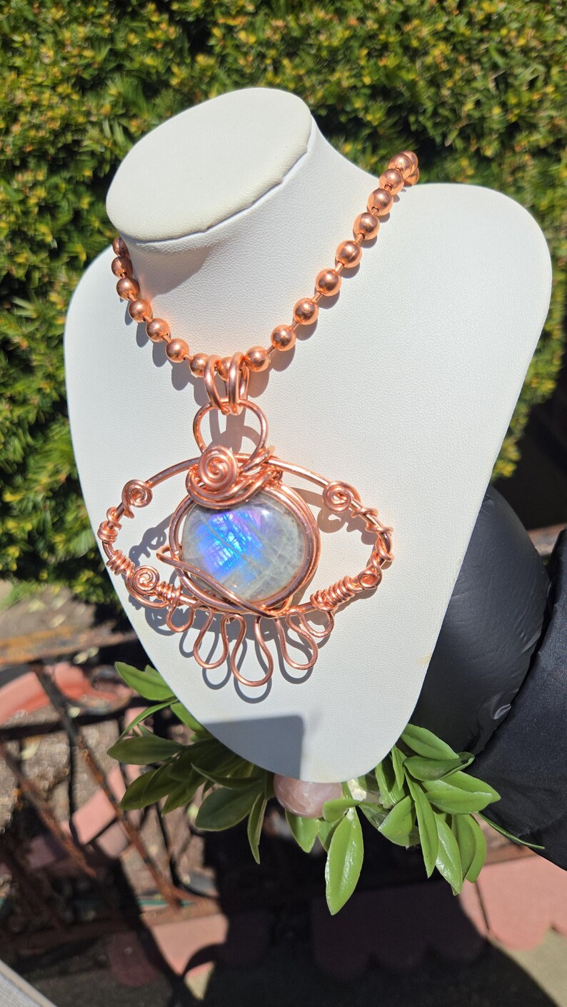Op de afbeelding: Een handgemaakte koperdraad hanger ketting met een grote, iriserende maansteen. De maansteen is gezet in een decoratief draadframe, hangend aan een koperen kogelketting. De hanger wordt getoond op een witte buste.