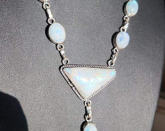 May include: A silver necklace with a triangular pendant featuring a white iridescent gemstone. The necklace has five round white gemstones.