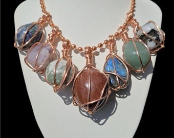 May include: A copper wire-wrapped necklace featuring seven different gemstones. The stones vary in color, including blue, purple, green, and brown. The necklace is displayed on a white mannequin bust.