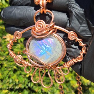 Op de afbeelding: Een handgemaakte hangerketting van koperdraad. De hanger heeft een grote, ovaalvormige, iriserende edelsteen, ingewikkeld gewikkeld in koperdraad. De ketting heeft een koperen kralenketting.
