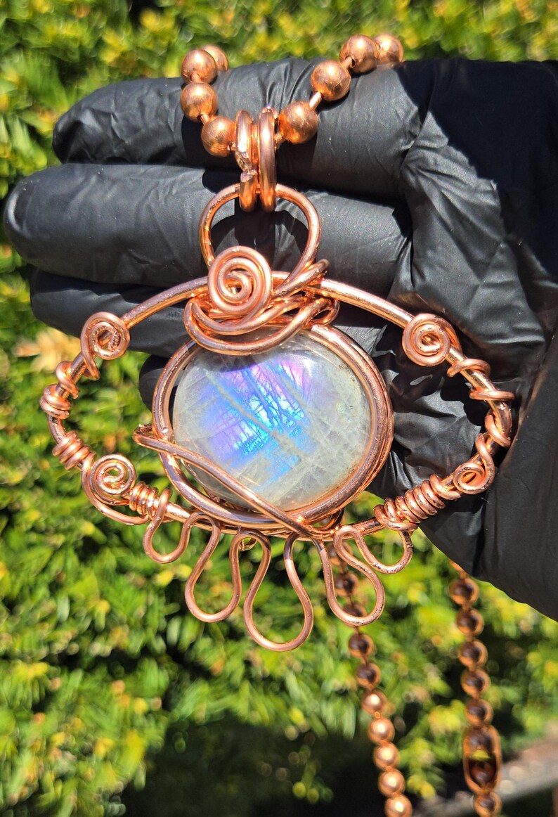 Op de afbeelding: Een handgemaakte hangerketting van koperdraad. De hanger heeft een grote, ovaalvormige, iriserende edelsteen, ingewikkeld gewikkeld in koperdraad. De ketting heeft een koperen kralenketting.