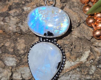 May include: A pendant featuring two moonstones set in silver-toned metal. The top stone is oval-shaped with blue iridescence, while the bottom stone is pear-shaped with a smooth, milky white surface. The setting has a beaded edge detail.