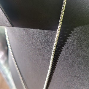 May include: A silver chain necklace with a box chain design. The chain is lying on a black surface.