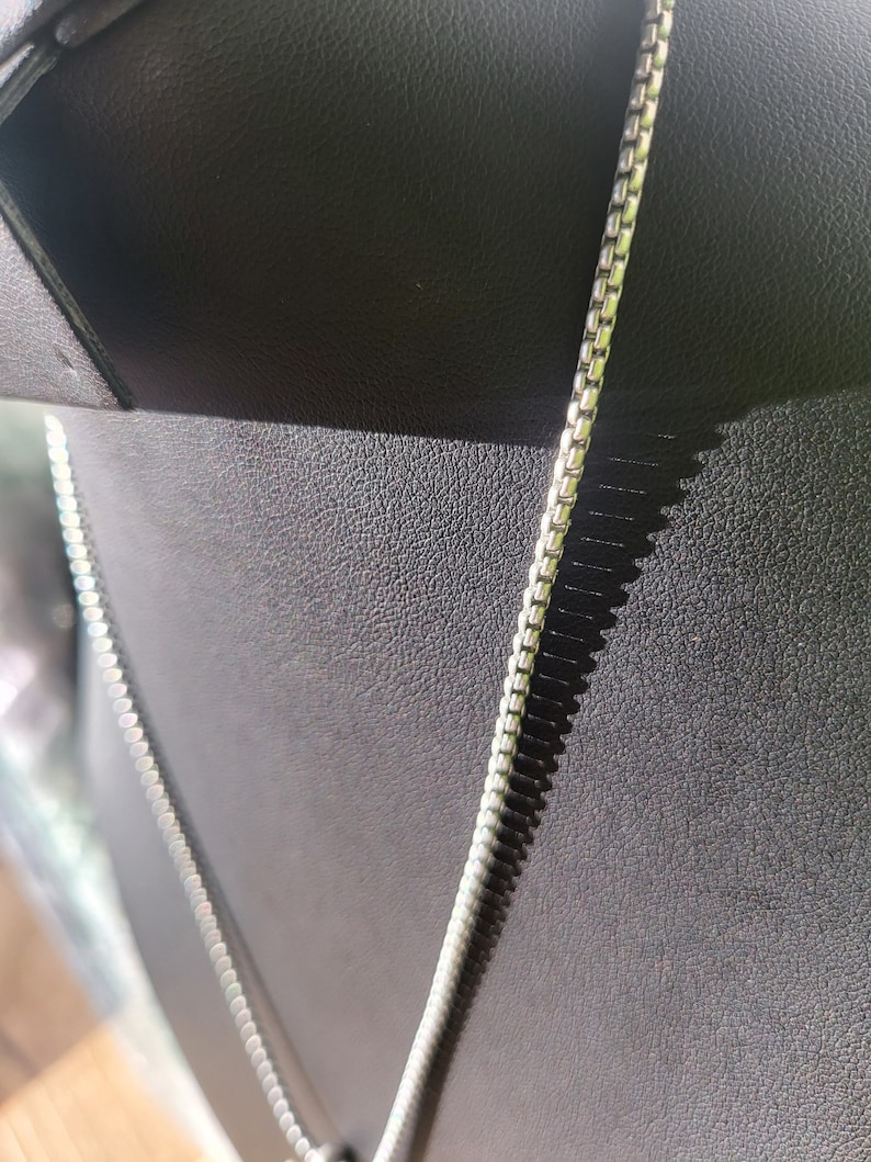 May include: A silver chain necklace with a box chain design. The chain is lying on a black surface.