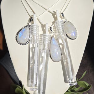 May include: A collection of three moonstone pendants and two clear plastic bubble wands suspended on white cords. The moonstones feature iridescent blue flashes and are set in silver-toned metal frames. The bubble wands are transparent cylinders.