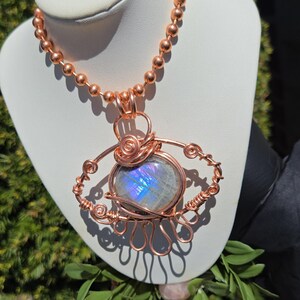 Op de afbeelding: Een koperdraad-gewikkelde ketting met een grote, ovale, iriserende edelsteen hanger. De hanger is het middelpunt, met ingewikkelde draaddetails rond de steen. De ketting is gemaakt van kleine koperen kralen.