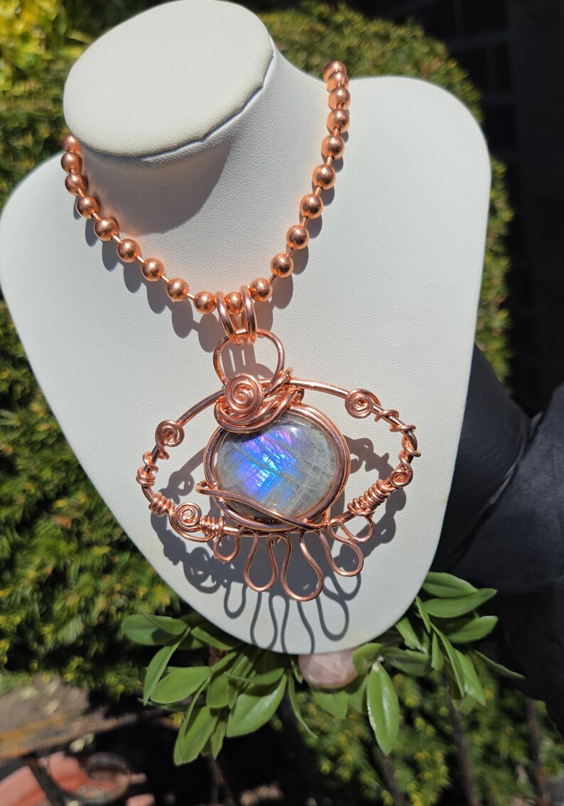 Op de afbeelding: Een koperdraad-gewikkelde ketting met een grote, ovale, iriserende edelsteen hanger. De hanger is het middelpunt, met ingewikkelde draaddetails rond de steen. De ketting is gemaakt van kleine koperen kralen.