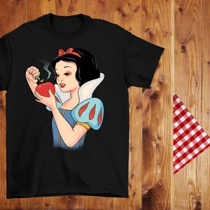 Może przedstawiać: Czarny t-shirt z kresk&oacute;wkową ilustracją Kr&oacute;lewny Śnieżki trzymającej dymiące czerwone jabłko. Ma na sobie niebiesko-białą sukienkę i czerwoną kokardę. Tło to drewniana powierzchnia z czerwoną i białą kraciastą szmatką.
