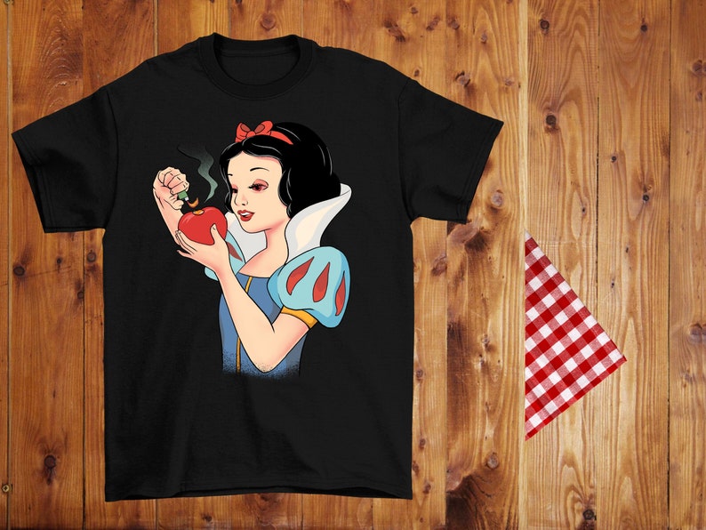 Może przedstawiać: Czarny t-shirt z kresk&oacute;wkową ilustracją Kr&oacute;lewny Śnieżki trzymającej dymiące czerwone jabłko. Ma na sobie niebiesko-białą sukienkę i czerwoną kokardę. Tło to drewniana powierzchnia z czerwoną i białą kraciastą szmatką.