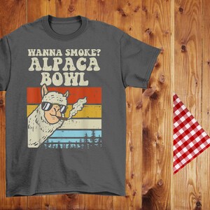 May include: A dark gray t-shirt with a vintage-style graphic of an alpaca wearing sunglasses and smoking a cigarette. The text "Wanna Smoke? Alpaca Bowl" is printed in white on the shirt.