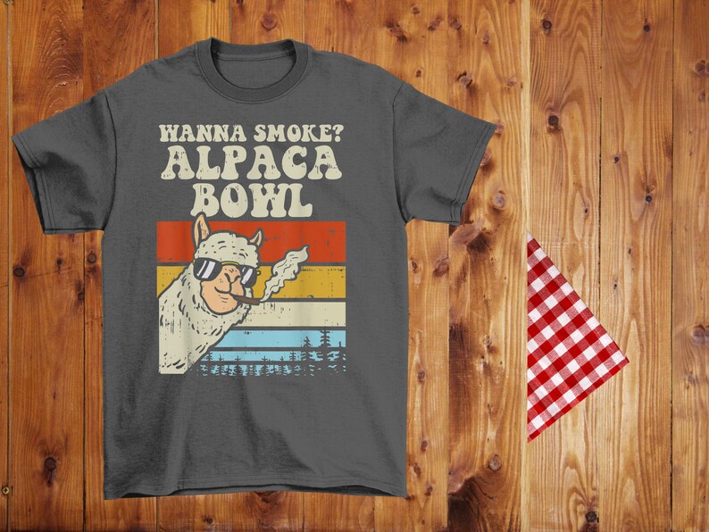 May include: A dark gray t-shirt with a vintage-style graphic of an alpaca wearing sunglasses and smoking a cigarette. The text "Wanna Smoke? Alpaca Bowl" is printed in white on the shirt.