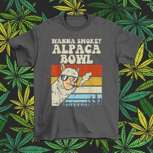 May include: A dark gray t-shirt with a retro-style graphic of an alpaca smoking a pipe. The text "Wanna Smoke? Alpaca Bowl" is printed in white on the shirt.