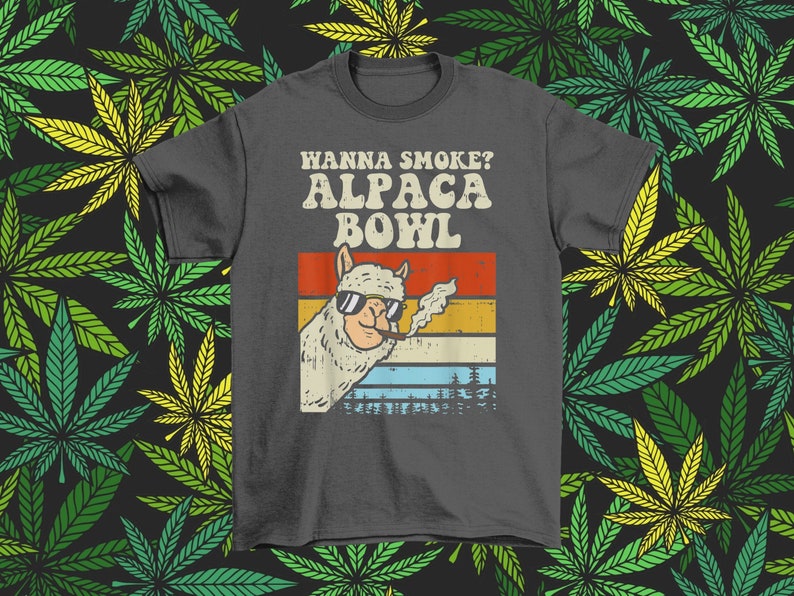 May include: A dark gray t-shirt with a retro-style graphic of an alpaca smoking a pipe. The text "Wanna Smoke? Alpaca Bowl" is printed in white on the shirt.