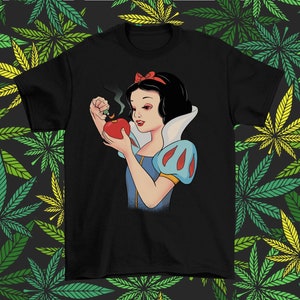 Może przedstawiać: Czarny t-shirt z kresk&oacute;wkową ilustracją Kr&oacute;lewny Śnieżki trzymającej czerwone jabłko, z kt&oacute;rego wydobywa się dym. Kr&oacute;lewna Śnieżka ma na sobie niebiesko-białą sukienkę i czerwoną kokardę. Tło to wz&oacute;r zielonych i ż&oacute;łtych liści konopi.