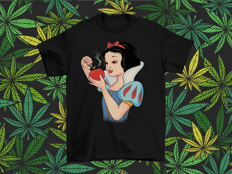 Może przedstawiać: Czarny t-shirt z kresk&oacute;wkową ilustracją Kr&oacute;lewny Śnieżki trzymającej czerwone jabłko, z kt&oacute;rego wydobywa się dym. Kr&oacute;lewna Śnieżka ma na sobie niebiesko-białą sukienkę i czerwoną kokardę. Tło to wz&oacute;r zielonych i ż&oacute;łtych liści konopi.