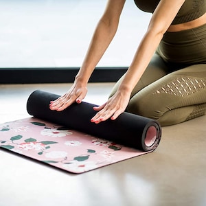 May include: A person rolls up a pink yoga mat with a floral pattern. The mat is black on the underside and has a pink edge. The person is wearing green leggings with cutouts and a matching sports bra.