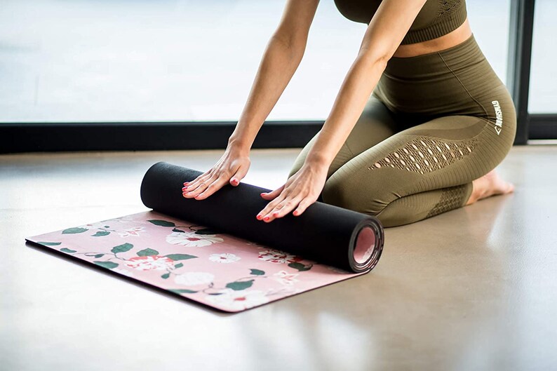 May include: A person rolls up a pink yoga mat with a floral pattern. The mat is black on the underside and has a pink edge. The person is wearing green leggings with cutouts and a matching sports bra.