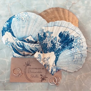 May include: Two white seashells with a blue and white wave design. The shells are arranged on a light brown surface with a small card that reads "Handmade by Periwinkle Shells".