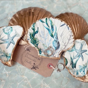 May include: Three gold-painted seashell ring dishes with a white background and blue and green floral designs. The shells are arranged in a triangle shape with a small tag that reads "Handmade by Periwinkle Shells".