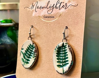 May include: A pair of ceramic earrings with a fern leaf design. The earrings are oval-shaped with a white background and a green fern leaf. The edges are outlined in a dark metallic color. The earrings are displayed on a brown card with the text "Moonlighter Ceramics".