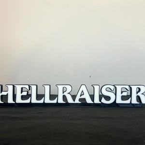 May include: A black and white sign that reads "HELLRAISER". The letters are white with a black outline, and the sign is on a dark surface. The sign is a decorative item.