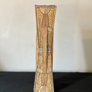 May include: A tall, rectangular, vertical object with a metallic gold finish and intricate, embossed geometric patterns resembling ancient hieroglyphics. The structure features symmetrical, raised detailing with subtle silver accents along the center.