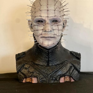 May include: A detailed bust of a character with a pale complexion, adorned with numerous metal pins embedded in the head. The figure wears a black textured garment with intricate detailing. The bust is set against a neutral background.