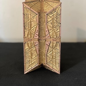 May include: A vertical, rectangular object with a metallic gold and copper finish, featuring intricate, geometric, and symmetrical relief patterns across its surface. The structure has a central vertical seam and angular, folded edges.