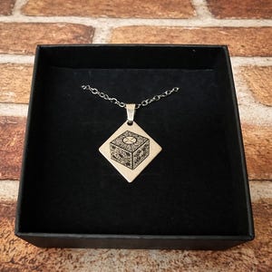 May include: A silver-colored necklace with a pendant in a black box. The pendant is a diamond shape with a detailed engraved cube design. The necklace chain is also silver. The box is black and sits on a brick background.