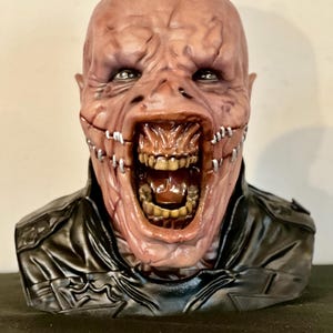 May include: A grotesque, detailed mask with a scarred, fleshy face, open mouth revealing teeth, and metal staples. The mask features a black leather-like jacket. The overall appearance is unsettling and horror-themed.